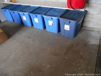 Six blue plastic bins arranged in a row containing Christmas decorations.