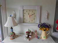 Wide shot showing clock on wall, lamp, pitcher with flowers, woman figurine, flower vase, and glass flower ornament on table surface beneath clock.