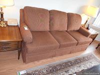 Front view of brown upholstered Ashley Furniture sofa with three seat cushions, placed on hardwood floor next to wooden side table and lamp.