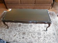 Full view of wooden coffee table with glass top and curved legs on a patterned area rug.