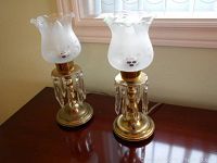 Pair of brass electric lamps with frosted glass flower-etched glass shades and crystal pendants, on a wooden surface near a window.