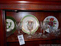 Three plates with floral and green decorative patterns, six crystal-cut style wine glasses, a small cut-glass ashtray, and a crystal-cut bowl displayed on a wooden shelf.