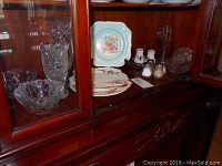 Display of assorted plates, crystal items, and condiment shakers arranged inside a wood cabinet.