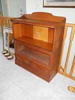 Full front view of the oak barrister's bookcase showing the open top shelf, missing glass door middle shelf, and bottom drawers, placed on a tiled floor next to artwork.