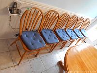 Six wooden dining chairs lined up against a wall. Chairs have rounded backs with vertical slats and blue tufted cushions on the seats. Light wood finish and tiled floor surrounding.