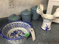 Photo showing three blue ceramic canisters with lids, the utensil holder with utensils inside, and the ceramic basket bowl on a kitchen counter.