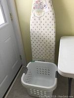 Ironing board with floral cover hanging against wall next to window and table, one square gray plastic laundry basket on floor nearby.