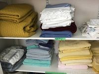 Two shelves filled with assorted linens including yellow quilted bedspreads, white pillow covers with lace, blankets, towels, doilies, and tablecloths