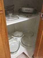Three Corningware covered casseroles with blue cornflower pattern, Pyrex and clear glass casserole dishes inside a white cabinet.