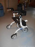 Side view of white Drive Nitro walker showing black padded seat, hand brakes, and three wheels (two smaller front wheels, one large rear wheel).