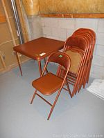 Photo showing the brown vinyl covered card table beside a stack of eight matching brown vinyl padded folding chairs in metal frames on a gray concrete floor against unfinished basement walls.