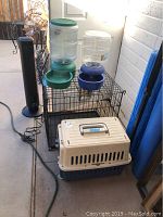 Wide view showing black wire pet pen, beige and navy blue pet carrier, and two self-watering pet bowls stacked on top of pen