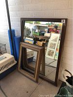 Both framed mirrors leaning against a wall, large mirror with plain dark brown frame and smaller mirror with decorative textured wooden frame in front.