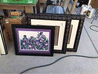 Three framed prints leaning against a wall. Front is a floral purple flower print with black frame and purple matting, two larger dark ornate framed prints behind.