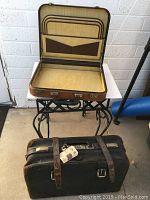 Open tan interior vintage briefcase with brown pocket inside the lid, resting on a metal stand. A closed dark brown vintage briefcase with leather straps and a handle is seen below on the floor.