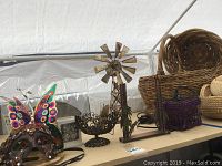 Wide view of metal windmill music box, Mardi Gras metal mask with butterfly design, curtain metal bowl, purple beaded hanging lamp, and woven baskets on table.