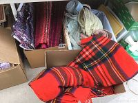 Three boxes containing fabric items. First box with thick woven rugs. Second box with folded towels and sheets. Third box with red plaid blanket and other fabric items.