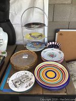 A collection of various plates including colorful striped plates, gold trimmed glass plates, hand-painted decorative ceramic plates on a metal stand, and ceramic plates with floral designs.