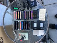 22 black plastic game/program cartridges on a round glass table with cords and an instruction sheet, plus Suncom joystick with white base and red button.