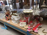 Photo of varied Christmas decor and kitchenware items including ceramic moose cookie jar, figurines, mugs, and flat gingerbread men ornaments on a wooden table.