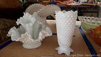Two white milk glass vases on table. One hobnail pattern vase with ruffled top, the other bud style vase with three separate ruffled bud holders.
