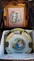 Framed ceramic wall hanging above and two decorative tin plates below in box