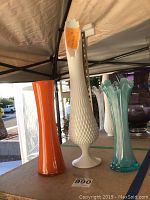 Photo shows three vases together on a table under canopy: white hobnail Fenton vase tallest, orange case glass vase medium height, and blue handmade glass vase shortest.