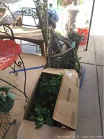 Box of assorted faux flowers with green leaves sitting on the floor, partially open box.