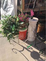 Wide view of the white decorative pedestal next to metal plant stand holding faux flowers, showing overall condition and size comparison to nearby objects