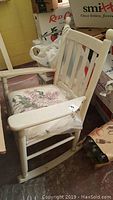 Wooden rocker chair with vertical back slats, padded floral seat, and worn cream paint. Partial view of room showing boxes and items around chair.