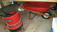 Red wheelbarrow with wooden handles and one white pneumatic tire positioned on a concrete floor next to red and black Vornado leaf eater.