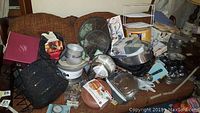 Wide shot of assorted pots, pans, and kitchen tools piled on chair including a food scale, tortilla warmer, and other kitchen items mostly in boxes or packaging.