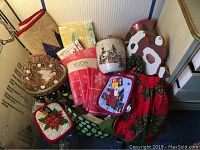 Wide view showing Christmas linens, ceramic bowls, pot holders, kitchen rugs and holiday decorations arranged in a box.