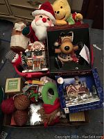 Two boxes on floor containing Santa plush, Winnie the Pooh plush, fiber optic reindeer in box, porcelain village set box, Christmas balls, gnome figure and some books.