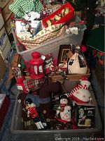 A plastic tote filled with Christmas decorations including plush reindeer, a red lantern candle holder, fabric snowman figurines, nutcracker, and assorted ornaments.