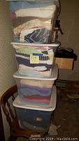 Four large plastic bins stacked on a wooden chair full of folded women's clothing with varied patterns and colors.