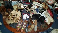 Image showing a large assortment of women's shoes carelessly arranged on a wooden round table, including boots, sandals, flats, slippers, and tennis shoes in neutral tones, black, and plaid patterns.
