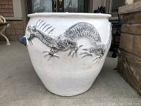 Front and side view of large white ceramic planter with detailed raised silver dragon design and two gray handles. Set on concrete floor outside a house.