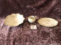 Three porcelain pieces on fabric backdrop: scalloped serving bowl, oval serving platter, cream pitcher, all decorated with gold gilt and floral patterns.