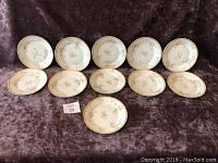 Full view of all 11 Limoges sandwich plates arranged on a dark fabric showing the full set and their floral design with gold gilt borders.