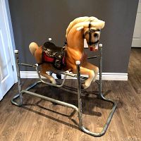 Front and side view showing the molded plastic horse with decorative black and red saddle, mounted on a metal spring frame with wear visible.