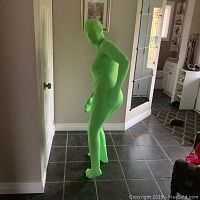 Person modeling bright green full-body chromakey suit including head, hands, and feet, standing sideways indoors