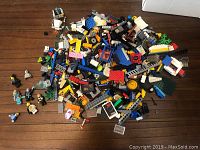 Image showing assorted LEGO pieces including bricks, minifigures, wheels, and special pieces on wooden floor.