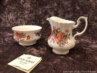 Royal Albert Centennial Rose bone china cream pitcher and sugar bowl with floral pattern and gold trim, displayed against a dark textured background, with Royal Albert tags.