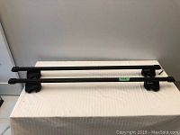 Pair of black GM roof racks with mounting brackets placed on a white surface.