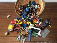 Basket filled with assorted LEGO pieces including bricks, plates, wheels, and a minifigure on a wooden floor