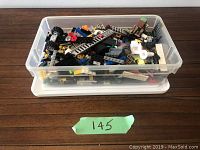 Clear plastic box filled with assorted LEGO pieces including wheels, bricks, ladders, and other elements
