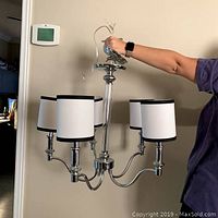 Front view of 5-light chandelier held by person, shows chrome finish and white shades with black trim.