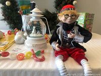 Photo of elf doll sitting with snow globe lantern on a white table near holiday decorations
