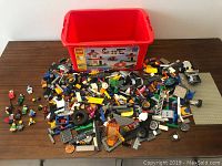 Photo showing red LEGO storage bin and large assortment of LEGO pieces spread out on a wooden surface, includes minifigures, wheels, bricks, ladders.